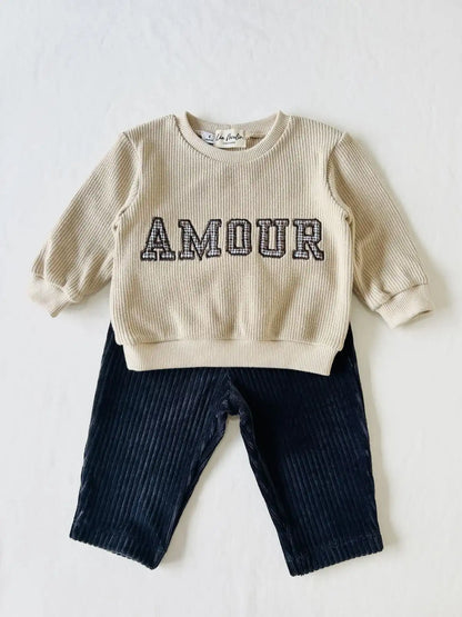 Inspiration de tenue bébé : pull léger beige brodé Amour associé à un pantalon souple en velours côtelé foncé.