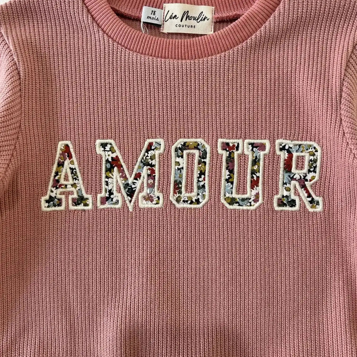 Gros plan sur la broderie Amour en tissu Liberty fleuri du pull bébé vieux rose