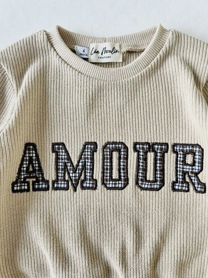 Gros plan sur la broderie vintage Amour avec motif à carreaux et le col extensible du pull bébé en maille côtelée.